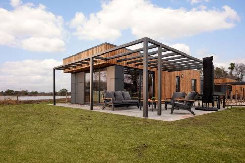 Moderne lodge met houten lambrisering, ruim terras en tuinmeubelen op een grasveld onder een blauwe lucht.