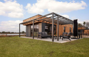 Lodge moderno con fachada de madera, amplia terraza y muebles de exterior sobre césped bajo cielo azul.