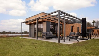 Moderne lodge med træbeklædning, stor terrasse og havemøbler på en græsplæne under blå himmel.