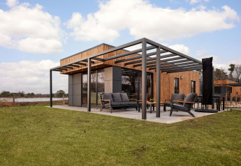 Modern lodge with wood cladding, large terrace and outdoor furniture, set on a grassy lawn under blue sky.