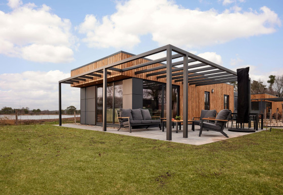 Moderne lodge med træbeklædning, stor terrasse og havemøbler på en græsplæne under blå himmel.