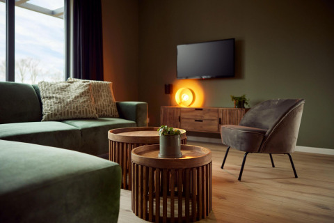 Salon moderne de lodge avec canapé vert, fauteuil, tables rondes en bois, TV et lumière ambiante chaleureuse.