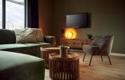 Sala de estar moderna en lodge con sofá verde, silla gris, mesas redondas, TV y luz cálida ambiental.