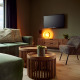 Soggiorno moderno in lodge con divano verde, poltrona grigia, tavolini rotondi, TV e luce calda e accogliente.
