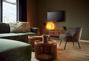 Soggiorno moderno in lodge con divano verde, poltrona grigia, tavolini rotondi, TV e luce calda e accogliente.