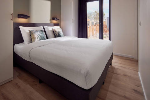 Bright, modern lodge bedroom with large bed, leaf-patterned pillows, wood floor, and window view outside.