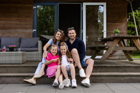 Familia sonriente sentada en las escaleras frente a su tienda Safarilodge en Camping Noetselerberg, Países Bajos.