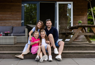 Familia sonriente sentada en las escaleras frente a su tienda Safarilodge en Camping Noetselerberg, Países Bajos.