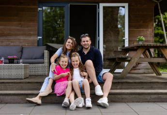 Familia sonriente sentada en las escaleras frente a su tienda Safarilodge en Camping Noetselerberg, Países Bajos.