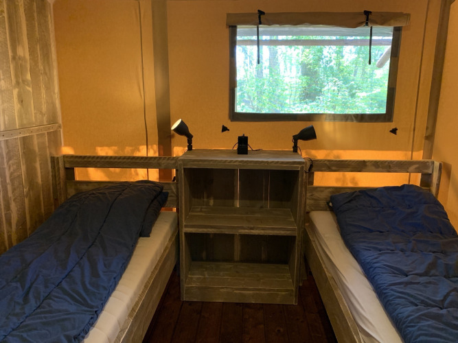 Two single beds with blue sleeping bags, shelf between them, and window with forest view at Glamphuys tent.