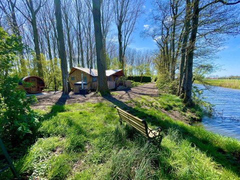 Tienda safari Glamphuys Ultra Wellness junto al agua en Buitengoed Ruysbos, Países Bajos, con árboles y sol.