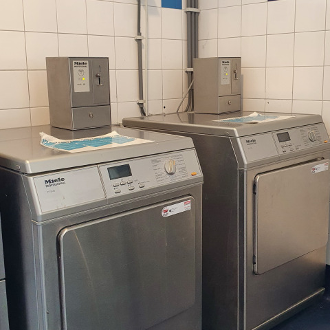 To professionelle Miele vaskemaskiner og tørretumblere i et glampingvaskeri med hvide fliser på væggene.