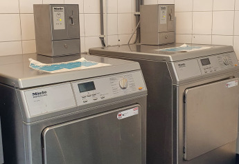 To professionelle Miele vaskemaskiner og tørretumblere i et glampingvaskeri med hvide fliser på væggene.