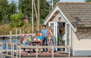 Famille profitant de la terrasse ensoleillée devant un chalet à Marina Parcs Almere, aux Pays-Bas, avec bateaux.