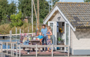 Family enjoying the sunny deck outside a chalet at Marina Parcs Almere, Netherlands, with boats behind.