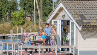 Familia disfruta una terraza soleada frente a un chalet en Marina Parcs Almere, Países Bajos, con barcos detrás.