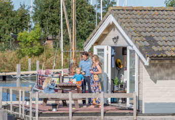 Family enjoying the sunny deck outside a chalet at Marina Parcs Almere, Netherlands, with boats behind.