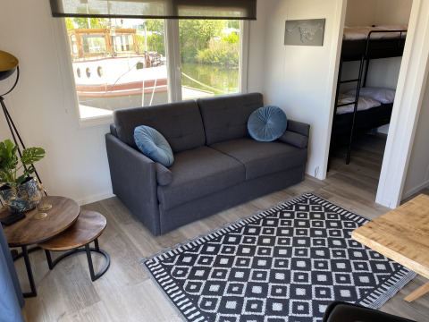 Stue med gråt sofa, blå puder, sort-hvidt tæppe, køjesenge og udsigt til båd ved Chalets at Marina Parcs Almere.
