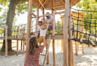 Gezin speelt samen op een houten speeltuig bij glampingverblijf, kinderen klimmen en lachen vrolijk.