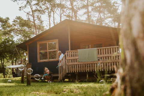 En familie nyder solen udenfor Youpi! Lodge, en safaritelt hytte omgivet af skov og store træer.