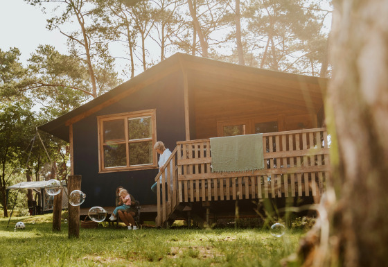 En familie nyder solen udenfor Youpi! Lodge, en safaritelt hytte omgivet af skov og store træer.