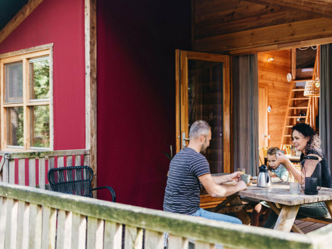 Familie sidder udenfor på terrassen ved et bord foran Youpi! Lodge safari telt på Beerze Bulten, Holland.