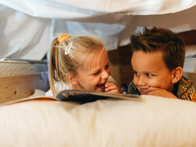 Two kids laughing and reading a book together under a blanket at Youpi! Lodge, Beerze Bulten, Netherlands.