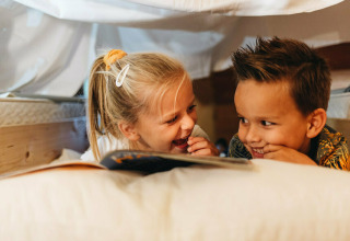 Twee kinderen lachen en lezen samen onder een deken in de Youpi! Lodge, Beerze Bulten, Nederland.