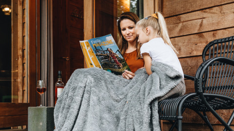 Madre e hija leen una revista bajo una manta frente a una tienda safari en Youpi! Lodge, Beerze Bulten.