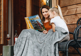 Madre e hija leen una revista bajo una manta frente a una tienda safari en Youpi! Lodge, Beerze Bulten.