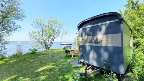 Lille blå hytte ved søen omgivet af træer, græsplæne og båd, Tiny Wagon ved Marina Parcs Almere, Holland.