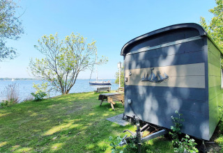 Small blue tiny house by the lake surrounded by trees and grass, Tiny Wagon at Marina Parcs Almere, Netherlands.