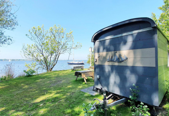 Lille blå hytte ved søen omgivet af træer, græsplæne og båd, Tiny Wagon ved Marina Parcs Almere, Holland.
