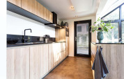 Cucina moderna di una tiny house con mobili in legno chiaro, rubinetto nero e grandi finestre luminose.