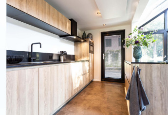 Cucina moderna di una tiny house con mobili in legno chiaro, rubinetto nero e grandi finestre luminose.