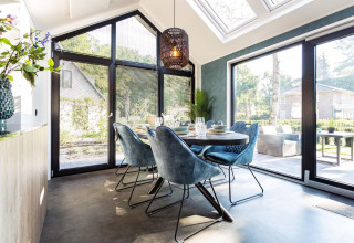 Zona pranzo luminosa e moderna in una tiny house con grandi finestre, sedie blu e vista sul giardino.