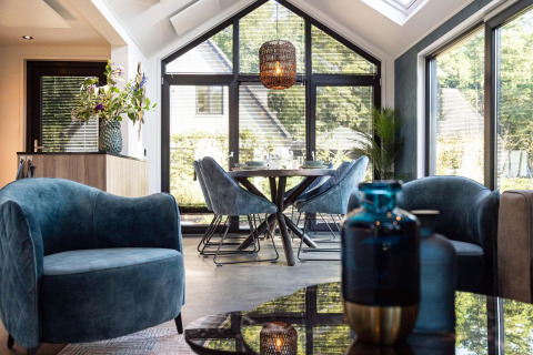 Moderne tiny house woonruimte met blauwe fluwelen stoelen, eettafel en grote ramen met zonlicht.