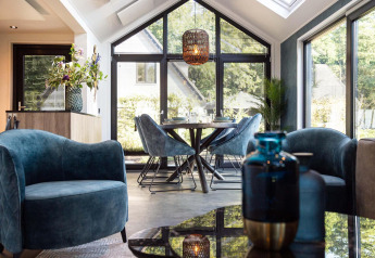 Zona giorno moderna di tiny house con poltrone blu, tavolo da pranzo e grandi finestre luminose.