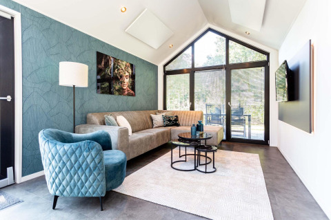 Moderne tiny house woonkamer met grote ramen, blauwe stoel, comfortabele bank en wanddecoratie.