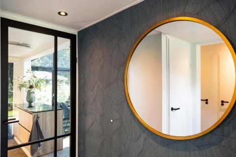 Intérieur moderne de tiny house avec un grand miroir rond, porte vitrée et cuisine ensoleillée avec fleurs.