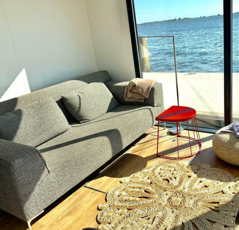 Soleado salón con sofá, mesa triangular roja y vista al agua desde la Houseboat 'Earrebarre' en Friesland, Países Bajos.