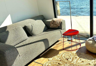 Soleado salón con sofá, mesa triangular roja y vista al agua desde la Houseboat 'Earrebarre' en Friesland, Países Bajos.