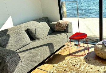 Soleado salón con sofá, mesa triangular roja y vista al agua desde la Houseboat 'Earrebarre' en Friesland, Países Bajos.