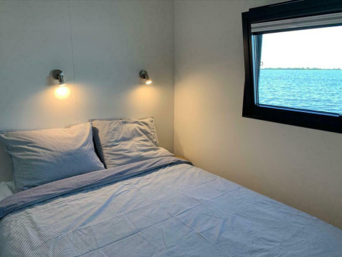 Camera da letto con letto matrimoniale, luci a parete e finestra con vista lago su una houseboat in Friesland.