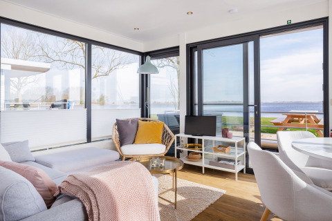 Soleado interior moderno de casa flotante con grandes ventanales, vista al agua y zona de estar en Friesland.