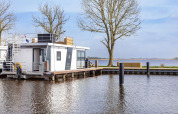 Modern houseboat on a peaceful lake in Friesland, Netherlands, with trees and outdoor seating nearby.