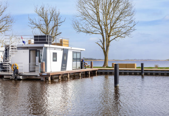 Modern houseboat on a peaceful lake in Friesland, Netherlands, with trees and outdoor seating nearby.