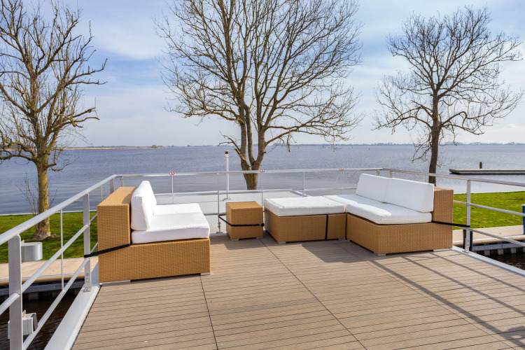 Outdoor lounge seating on a houseboat in Friesland, Netherlands, overlooking a lake with bare trees.