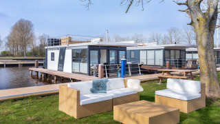 Udendørs siddeområde nær træ, med moderne husbåd på vandet i Friesland, Holland, under blå himmel.