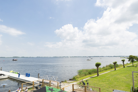 Vista desde una casa flotante hacia un lago con barcos, muelle, césped verde y cielo parcialmente nublado.